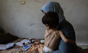 Palestinian-Mother-blue-headwrap-cradling-starving-child-counting-few-pills-in-hand